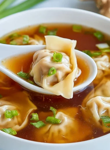 A bowl of steaming wonton soup garnished with green onions and herbs.